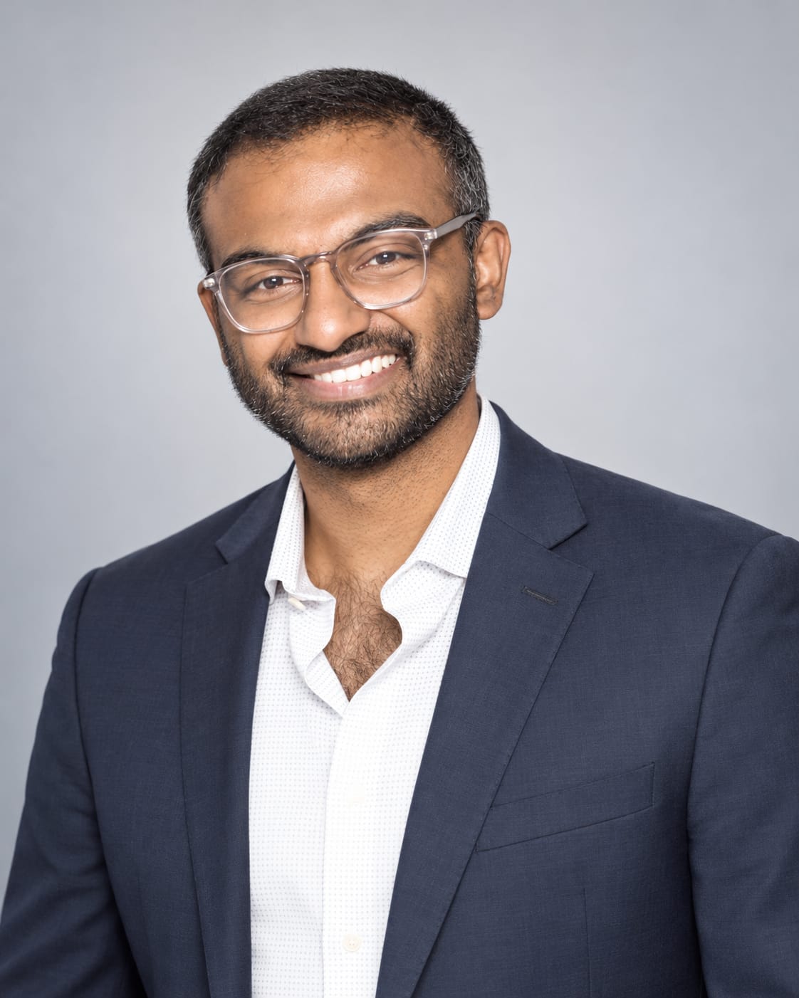 Jordan Pereira smiling, wearing glasses and a navy suit jacket
